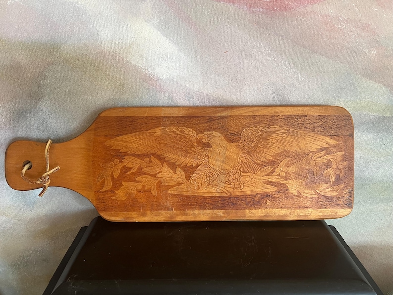 May include: A rectangular wooden serving board with a handle featuring a hole and a leather tie. The surface displays a detailed laser-engraved image of a bald eagle with wings spread wide, perched above a branch with leaves and berries.