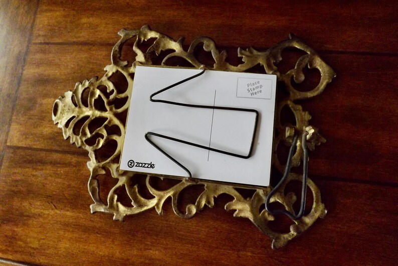 May include: Ornate gold-colored metal postcard holder with a white postcard. The postcard has a black wire design and the text "Place Stamp Here". The Zazzle logo is visible on the postcard.