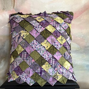 May include: A square throw pillow featuring a textured, layered design of folded fabric triangles. The surface is composed of a repeating pattern of purple, lavender, and olive green fabric squares with various floral and plaid prints.