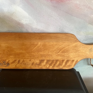 May include: A rectangular wooden serving board with a smooth, light brown finish and a handle on the right side. A leather cord is looped through a hole in the handle. The board features a small oval stamp that reads 1854 THE OLD DRUM SHOP GRANVILLE, MASS.