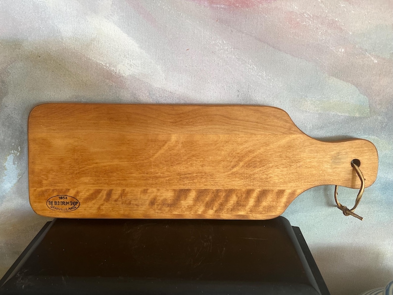 May include: A rectangular wooden serving board with a smooth, light brown finish and a handle on the right side. A leather cord is looped through a hole in the handle. The board features a small oval stamp that reads 1854 THE OLD DRUM SHOP GRANVILLE, MASS.