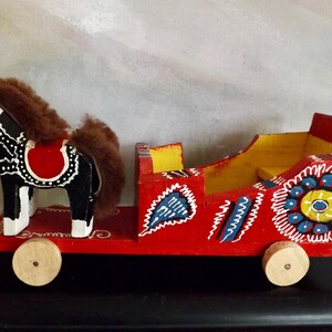 Puede incluir: Un juguete de madera de un caballo y un carruaje pintados en rojo, negro, blanco, amarillo, azul y verde. El caballo tiene una melena y una cola marrones. El carruaje tiene ruedas y un asiento.