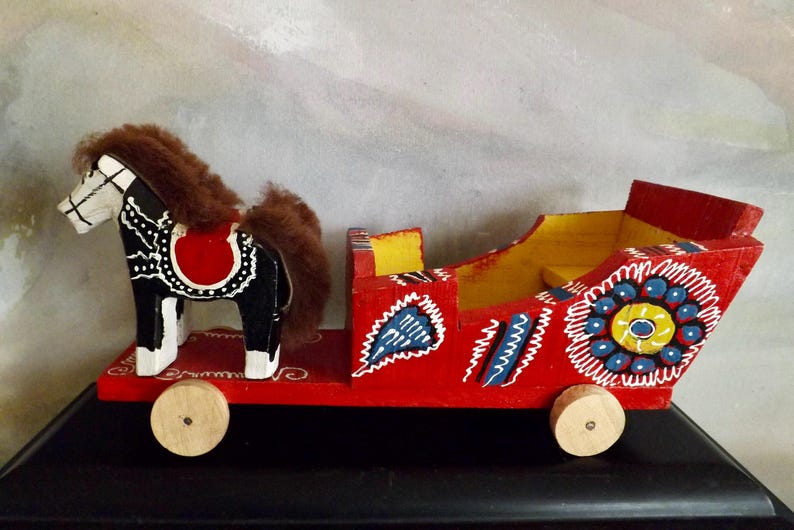 Puede incluir: Un juguete de madera de un caballo y un carruaje pintados en rojo, negro, blanco, amarillo, azul y verde. El caballo tiene una melena y una cola marrones. El carruaje tiene ruedas y un asiento.