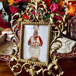 May include: Ornate gold-colored picture frame with a vintage Easter illustration. The illustration features a rabbit holding a tray of colorful Easter eggs. The frame is surrounded by a vibrant bouquet of flowers, creating a festive display.