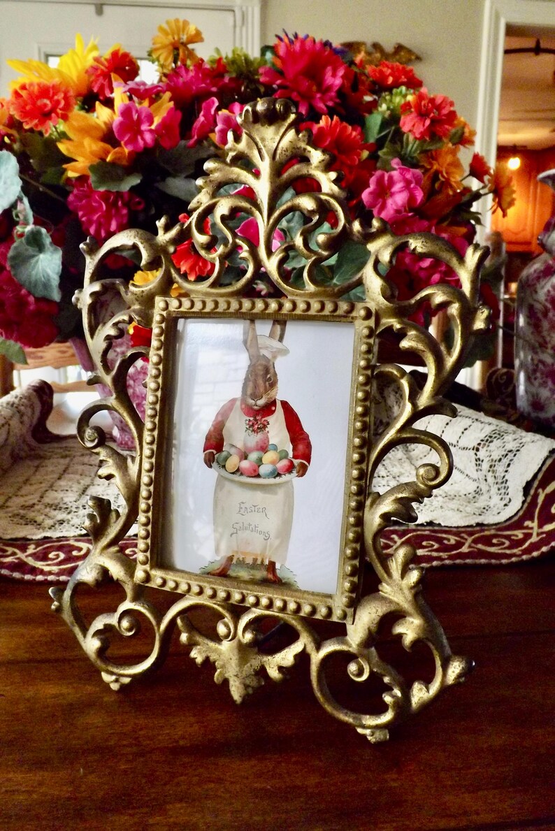 May include: Ornate gold-colored picture frame with a vintage Easter illustration. The illustration features a rabbit holding a tray of colorful Easter eggs. The frame is surrounded by a vibrant bouquet of flowers, creating a festive display.