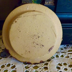 May include: A round, cream-colored ceramic plate with a raised rim and a slightly textured surface. The plate has a worn, vintage look.