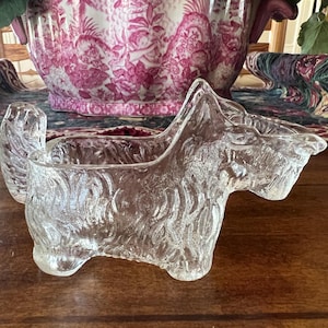May include: A clear glass figurine shaped like a Scottish Terrier dog, featuring a textured surface that mimics fur. The hollow body design allows it to function as a small dish or planter, with an upright tail and a distinct snout profile.