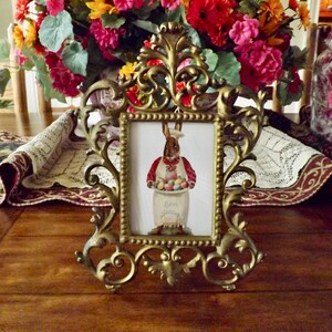 May include: Ornate gold-colored picture frame with a rectangular image of a bunny holding Easter eggs. The frame is on a wooden table, with a floral arrangement and decorative vases in the background.