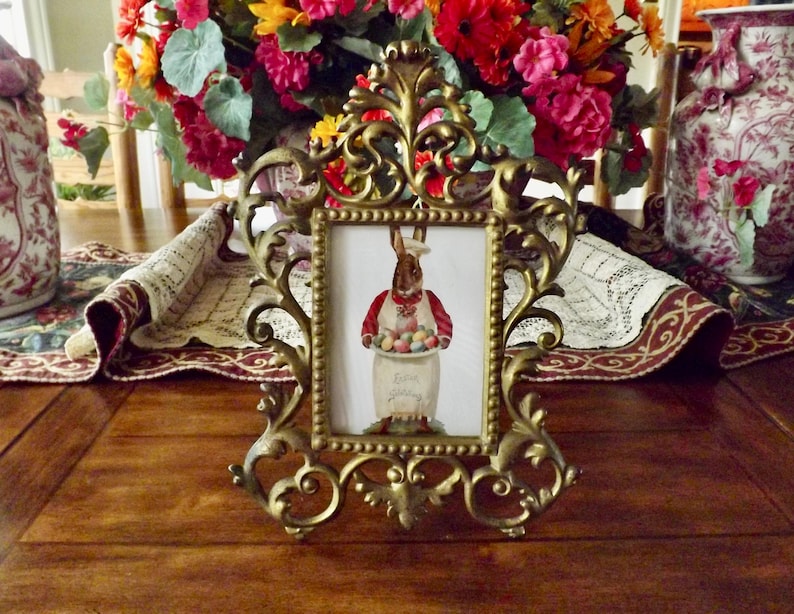 May include: Ornate gold-colored picture frame with a rectangular image of a bunny holding Easter eggs. The frame is on a wooden table, with a floral arrangement and decorative vases in the background.