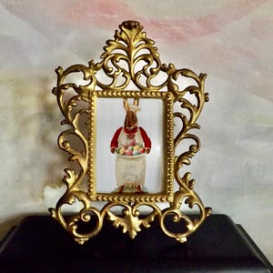 May include: Ornate gold-colored picture frame with a rectangular opening. The frame surrounds a picture of a rabbit wearing a red vest and holding a basket of Easter eggs. The words "Easter Spring" are visible.