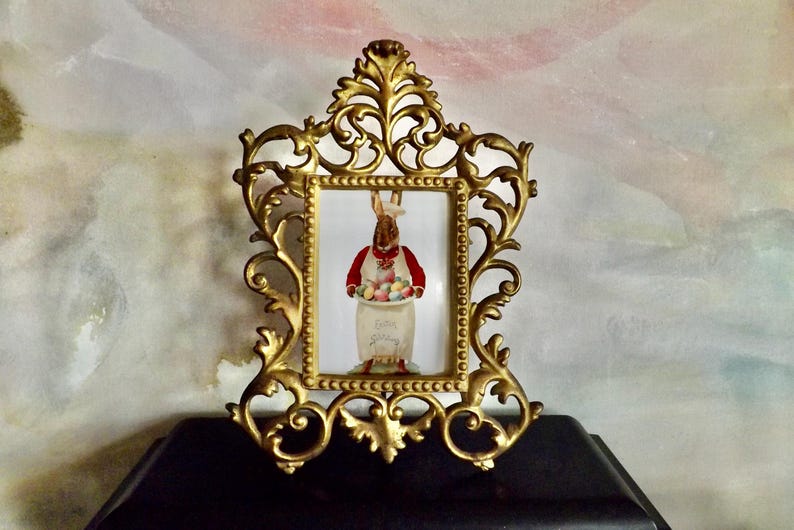 May include: Ornate gold-colored picture frame with a rectangular opening. The frame surrounds a picture of a rabbit wearing a red vest and holding a basket of Easter eggs. The words "Easter Spring" are visible.