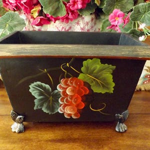 May include: Black planter with hand-painted grapes and leaves. The planter has three decorative feet and a distressed wood finish.