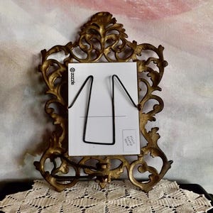 May include: Ornate, antique gold-colored picture frame with a white card and black wire holder. The frame has a detailed, scrolling design. The card has a black wire stand. The background is a watercolor-style painting.