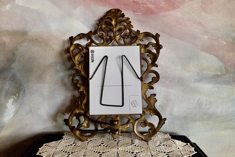 May include: Ornate, antique gold-colored picture frame with a white card and black wire holder. The frame has a detailed, scrolling design. The card has a black wire stand. The background is a watercolor-style painting.