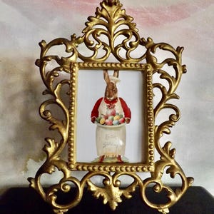 May include: Ornate gold-colored picture frame with a rectangular opening. Inside the frame is a picture of a brown rabbit wearing a chef's hat and apron, holding a jar filled with colorful Easter eggs. The jar has the words "Easter Greetings".