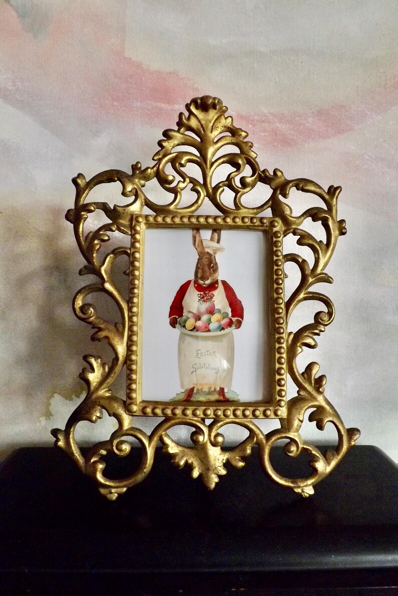 May include: Ornate gold-colored picture frame with a rectangular opening. Inside the frame is a picture of a brown rabbit wearing a chef's hat and apron, holding a jar filled with colorful Easter eggs. The jar has the words "Easter Greetings".