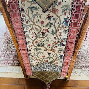 May include: A rectangular table runner featuring a central cream-colored panel with a woven pattern of trees, monkeys, and figures. It is bordered by wide, pink velvet strips with a geometric grid design and finished with a pointed end and tassel.