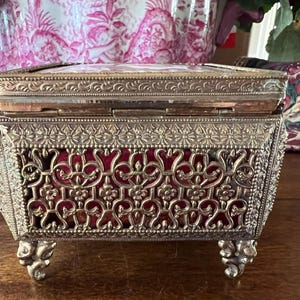 May include: A rectangular metal jewelry box featuring an intricate gold-tone filigree exterior with floral and scrollwork patterns. The box is lined with a deep red velvet interior and stands on four ornate, decorative metal feet with a hinged lid.