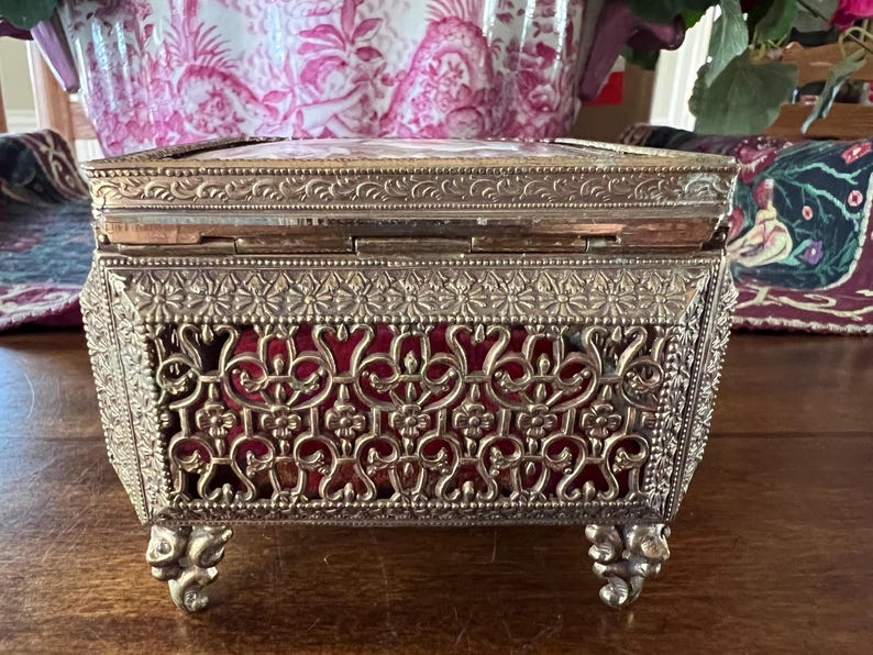 May include: A rectangular metal jewelry box featuring an intricate gold-tone filigree exterior with floral and scrollwork patterns. The box is lined with a deep red velvet interior and stands on four ornate, decorative metal feet with a hinged lid.