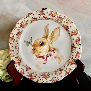 Puede incluir: Plato decorativo con una ilustración de un conejo y una mariposa. El plato tiene un borde floral con rosas rojas y rosas. El plato se exhibe sobre un soporte marrón oscuro.