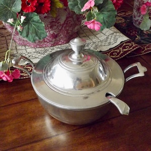 May include: A silver-colored serving bowl with a lid and a spoon. The bowl has two handles and a knob on the lid. The bowl is sitting on a wooden surface.
