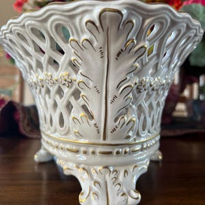 Puede incluir: Un cuenco de cer&aacute;mica blanca con un dise&ntilde;o de celos&iacute;a calada, con detalles de hojas pintadas en oro y peque&ntilde;as flores. El cuenco tiene un borde festoneado y se apoya sobre tres pies ornamentados, con bordes dorados que resaltan la base.