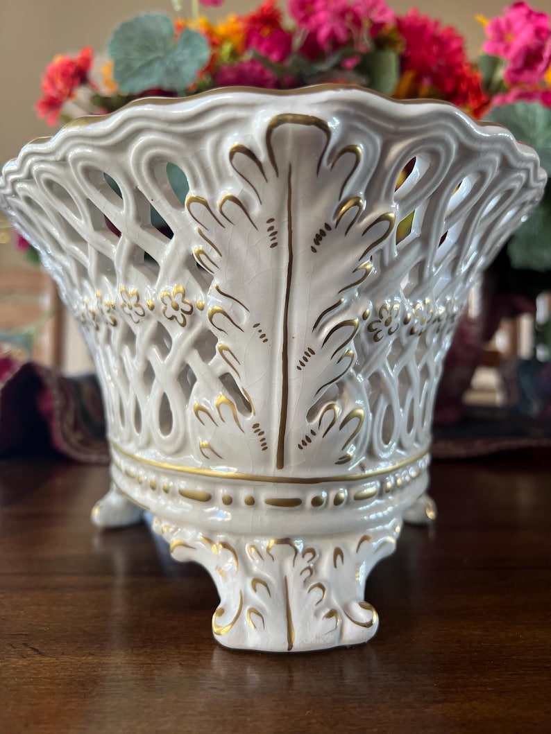 Puede incluir: Un cuenco de cer&aacute;mica blanca con un dise&ntilde;o de celos&iacute;a calada, con detalles de hojas pintadas en oro y peque&ntilde;as flores. El cuenco tiene un borde festoneado y se apoya sobre tres pies ornamentados, con bordes dorados que resaltan la base.