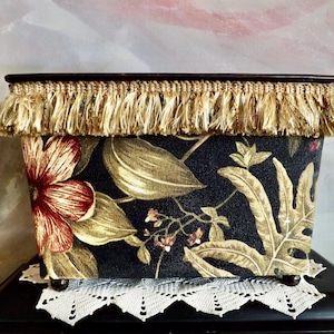 May include: A rectangular storage box with a floral pattern in red, gold, and green on a dark blue background. The top edge is trimmed with a brown border and gold fringe. The box sits on a white lace doily.