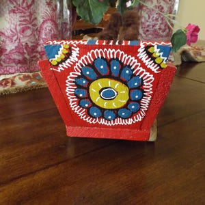 Puede incluir: Una caja de madera roja con un dise&ntilde;o pintado de flores azules, amarillas y blancas. La caja est&aacute; decorada con un borde blanco y tiene un estilo de arte popular tradicional.