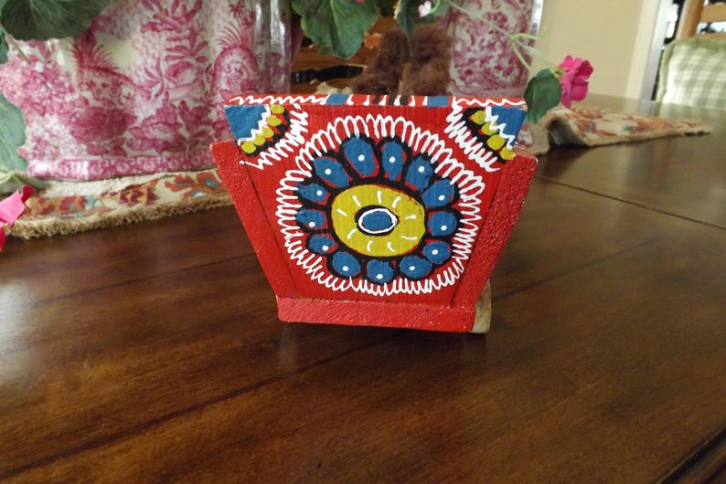 Puede incluir: Una caja de madera roja con un dise&ntilde;o pintado de flores azules, amarillas y blancas. La caja est&aacute; decorada con un borde blanco y tiene un estilo de arte popular tradicional.