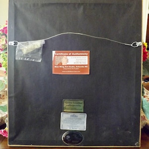 May include: The back of a framed artwork with a certificate of authenticity from Blue Wing Art Studio, Asheville NC. The certificate states that the artwork was prepared in the studio of the artist Jennifer Barrineau. The artwork is also labeled as "Quality Assembled and Inspected by Tri-Guard".