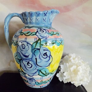 Puede incluir: Una jarra de cerámica azul y amarilla con un diseño floral con rosas azules. La jarra tiene una boca ancha y un asa.