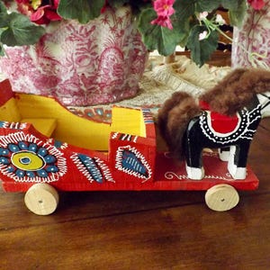 Puede incluir: Un carro de juguete de madera rojo, amarillo y azul tirado por un caballo negro con una crin marr&oacute;n. El carro tiene ruedas y est&aacute; decorado con patrones coloridos.