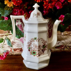 May include: White ceramic teapot with a floral design in brown and green. The teapot has a lid and a spout. It is sitting on a table with a floral tablecloth.