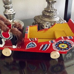 Puede incluir: Un juguete de madera de un caballo y un carruaje pintados en colores brillantes. El caballo es negro con marcas blancas y una silla de montar roja. El carruaje es rojo con detalles amarillos, azules y blancos. El juguete est&aacute; hecho a mano y tiene un encanto r&uacute;stico.