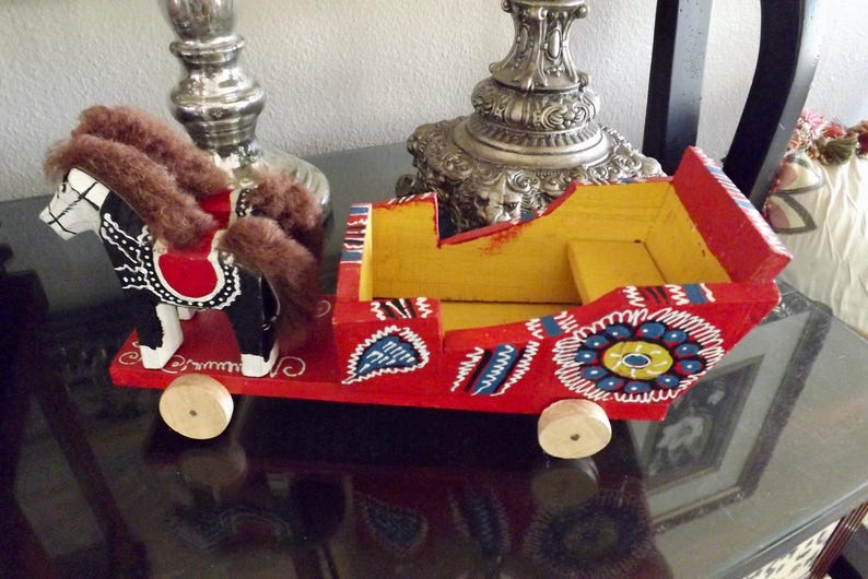 Puede incluir: Un juguete de madera de un caballo y un carruaje pintados en colores brillantes. El caballo es negro con marcas blancas y una silla de montar roja. El carruaje es rojo con detalles amarillos, azules y blancos. El juguete est&aacute; hecho a mano y tiene un encanto r&uacute;stico.