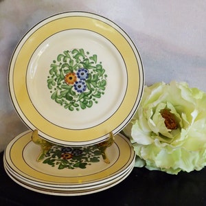 Puede incluir: Un juego de cuatro platos de cena de cerámica blanca con un borde amarillo y un diseño floral en el centro. El diseño floral presenta hojas verdes y flores azules, amarillas y naranjas.