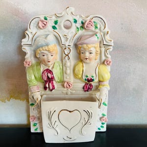 May include: A cream-colored ceramic wall pocket featuring two figures in period attire, one in a green jacket and one in a yellow top. The piece is decorated with pink roses, gold trim, and a central heart motif on the pocket, with a hanging hole.
