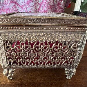 May include: A rectangular metal jewelry box with an ornate gold-tone finish, featuring intricate filigree cutouts, a beaded border, and four decorative feet. The interior is lined with deep red velvet, and the lid displays a floral design.