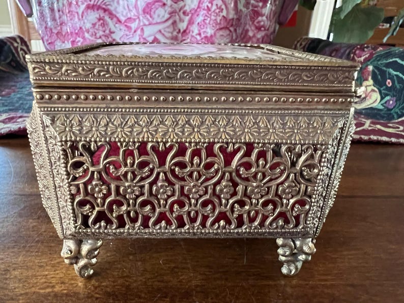 May include: A rectangular metal jewelry box with an ornate gold-tone finish, featuring intricate filigree cutouts, a beaded border, and four decorative feet. The interior is lined with deep red velvet, and the lid displays a floral design.