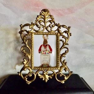 May include: Ornate gold-colored picture frame with a rectangular opening. Inside the frame is a picture of a bunny wearing a red vest and holding a jar filled with Easter eggs. The jar has the words "Easter Spring" on it.