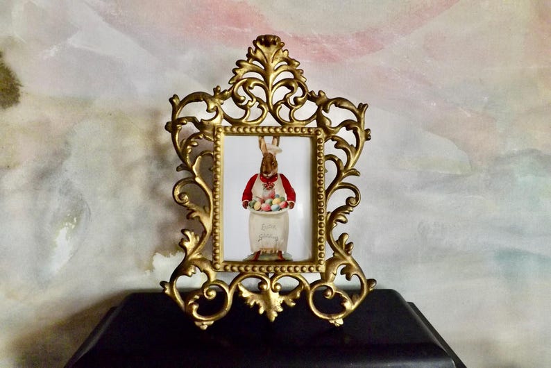 May include: Ornate gold-colored picture frame with a rectangular opening. Inside the frame is a picture of a bunny wearing a red vest and holding a jar filled with Easter eggs. The jar has the words "Easter Spring" on it.