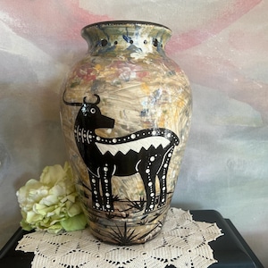 May include: A ceramic vase with a glossy, multi-colored marbled finish in shades of tan, cream, and blue. The vase features a prominent black silhouette of a four-legged animal with white dotted details and a jagged white stripe along its back.