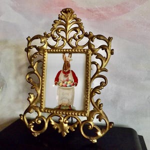 May include: Ornate gold-colored picture frame with a rectangular image of an Easter bunny holding a tray of colorful eggs. The bunny wears a red vest and chef's hat. The frame has a detailed, scrolling design.