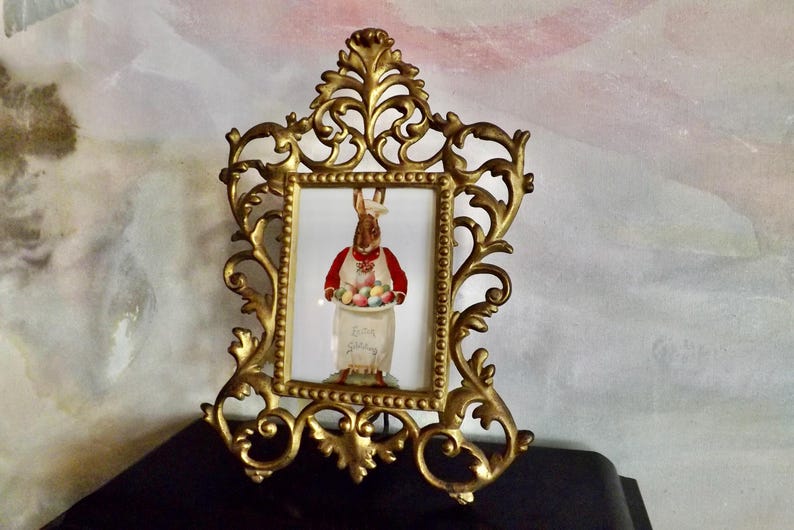May include: Ornate gold-colored picture frame with a rectangular image of an Easter bunny holding a tray of colorful eggs. The bunny wears a red vest and chef's hat. The frame has a detailed, scrolling design.