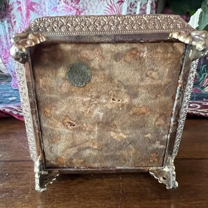May include: A square metal box with a textured gold-tone border and four ornate claw-style feet. The underside is covered in a tan, worn felt material and features a small, circular, dark-colored metal seal or emblem attached to the surface.