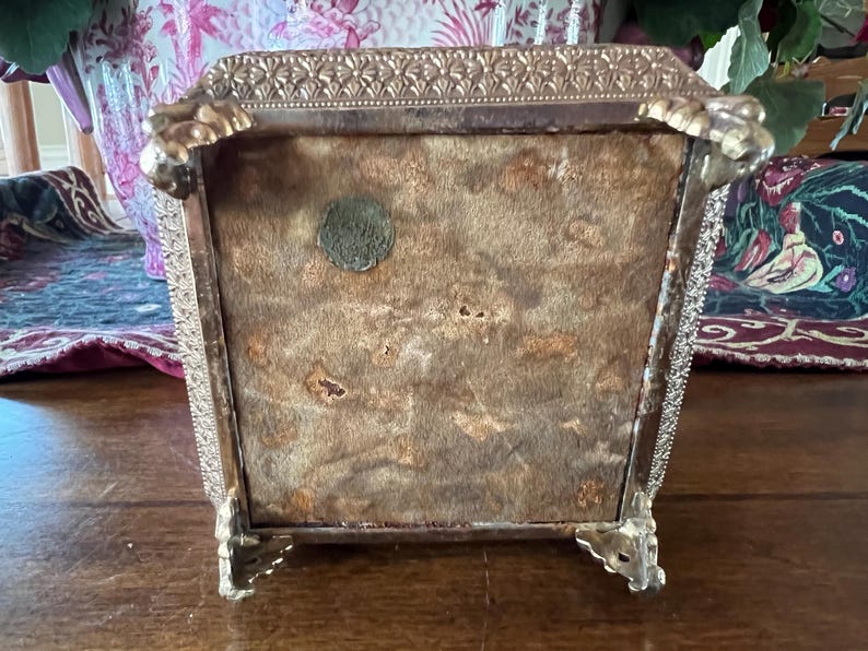 May include: A square metal box with a textured gold-tone border and four ornate claw-style feet. The underside is covered in a tan, worn felt material and features a small, circular, dark-colored metal seal or emblem attached to the surface.