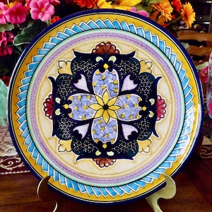 May include: A decorative ceramic plate with a vibrant floral design. The plate features a yellow center with a blue and white floral pattern, surrounded by a yellow, blue, and purple border. The plate is displayed on a gold-colored stand.