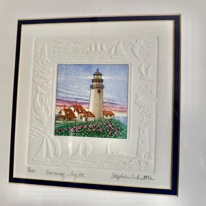 May include: Framed artwork featuring a lighthouse scene. The artwork depicts a tall, white lighthouse with a red-roofed building, set against a colorful sky. The frame is dark blue with a white embossed border of sailboats and lighthouses.