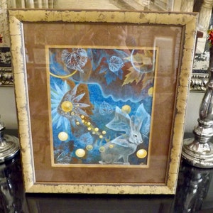 May include: A framed painting of a white rabbit with large ears, surrounded by blue and gold abstract shapes and leaves. The frame is brown with a gold trim.